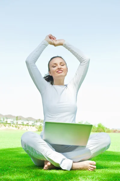Довольно расслабленная женщина, сидящая на траве и вытягивая руки, прицеливается в небо, работая над ноутбуком — стоковое фото