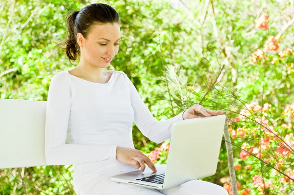 Junge hübsche Frau mit Laptop sitzt auf der Bank in einem Park — Stockfoto