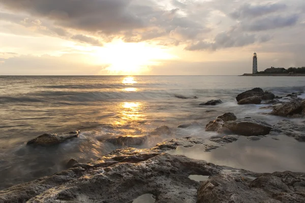 Krásné kamenné moře pláž při západu slunce s majákem — Stock fotografie