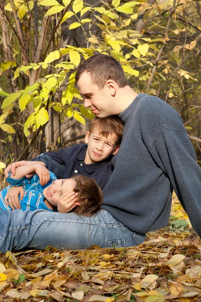 Отец и двое сыновей сидят на опавших листьях в парке — стоковое фото
