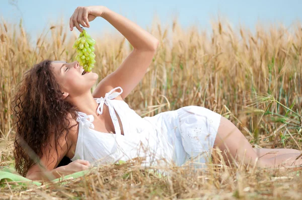 Dokonalá žena jíst hrozny v pšeničné pole. piknik. — Stock fotografie