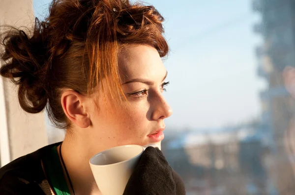 Красива молода жінка з чашкою кави над вікном — стокове фото