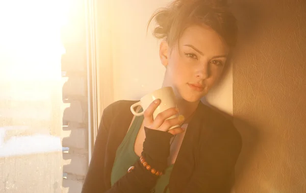 Vacker ung kvinna med kopp hett kaffe över solen fönster — Stockfoto