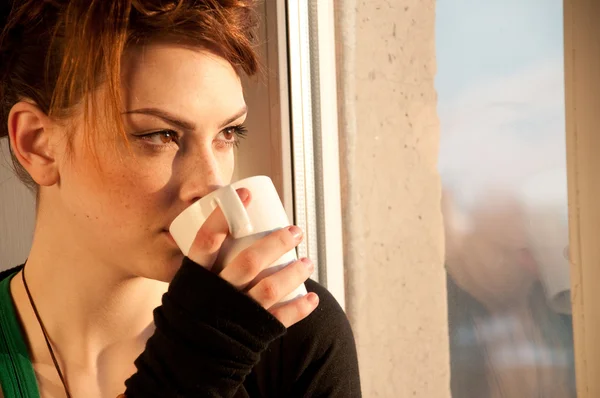 Красива молода жінка з чашкою кави над вікном — стокове фото