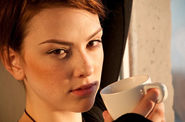 Mooie jonge vrouw met kop koffie over venster — Stockfoto