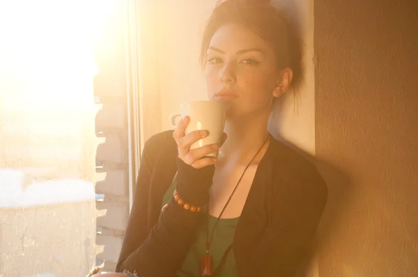 Красива молода жінка з чашкою гарячої кави над сонячним вікном — стокове фото