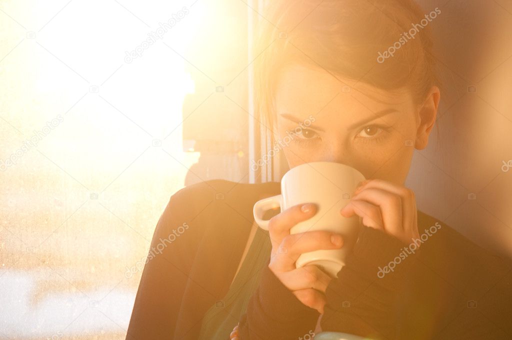 Beautiful young woman with cup of hot coffee over sun window