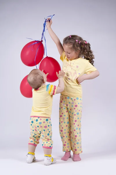 Twee leuke meisjes in roze pyjama's spelen met rode ballonnen — Stockfoto