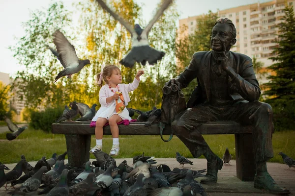 A little girl feeding the pigeons in the park near the statue — Stock Photo, Image