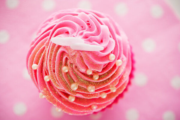 A cupcake in a pink baking cups with pink cream, white decoration and two hearts on the top on pink background as a studio shoot