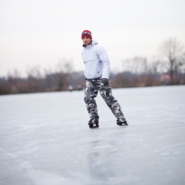 Handsome young man ice skating outdoors on a pond on a cloudy wi clipart