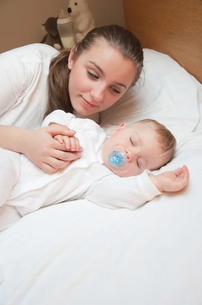 Happy young beautiful mother on the bed with her sleeping baby — Stock Photo, Image