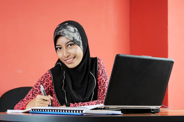 Schöne junge Muslimin lächelt, während sie vor dem Notizbuch schreibt — Stockfoto
