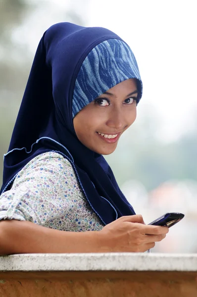 Schöne junge Muslimin lächelt, während sie Text auf dem Handy schreibt — Stockfoto