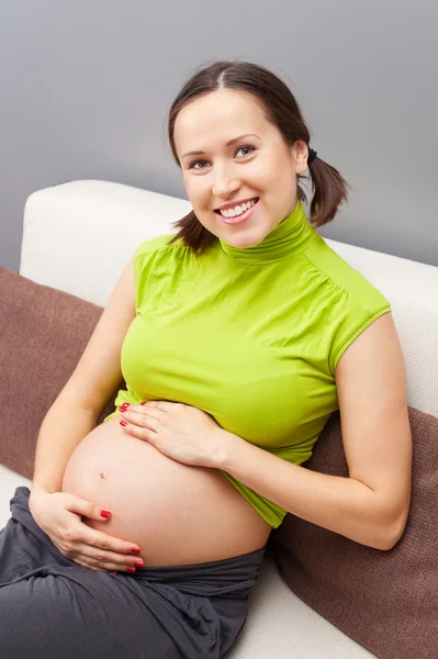 Terhes nő ölelés hasát, és ül a kanapén — Stock Fotó