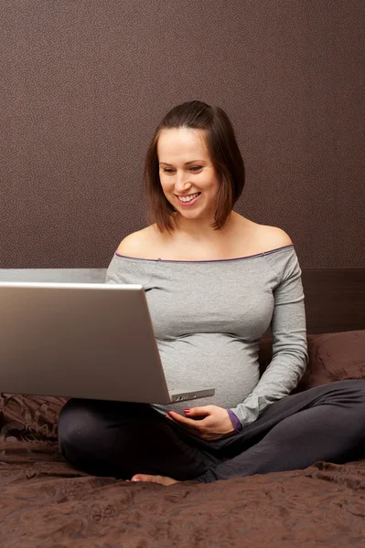 Беременная женщина с ноутбуком сидит на кровати — стоковое фото
