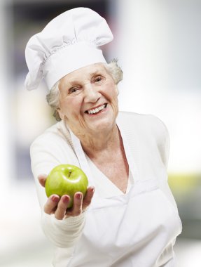 Senior woman cook offering a green apple, indoor