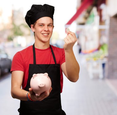 Portrait of young cook man holding euro coin and piggy bank at s clipart