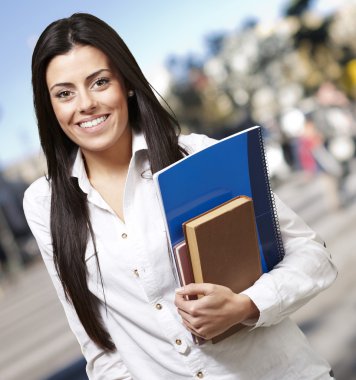 Pretty young woman smiling and holding notebooks against a stree clipart