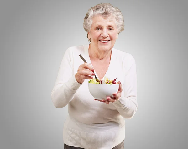 Ritratto di donna anziana che mangia insalata su sfondo grigio — Foto Stock