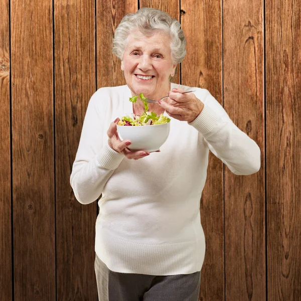 Portrait of senior woman eating a fresh salad against a wooden w — Stock Photo, Image
