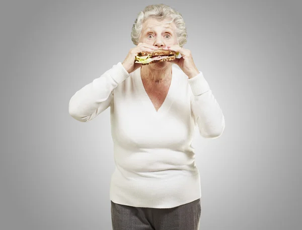 Mulher idosa comer um sanduíche saudável contra um fundo cinza — Fotografia de Stock