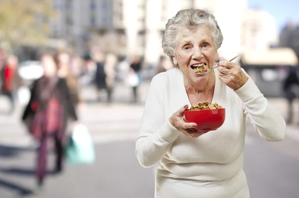 Senior woman eating cereals out of a red bowl against a street b — Stock Photo, Image