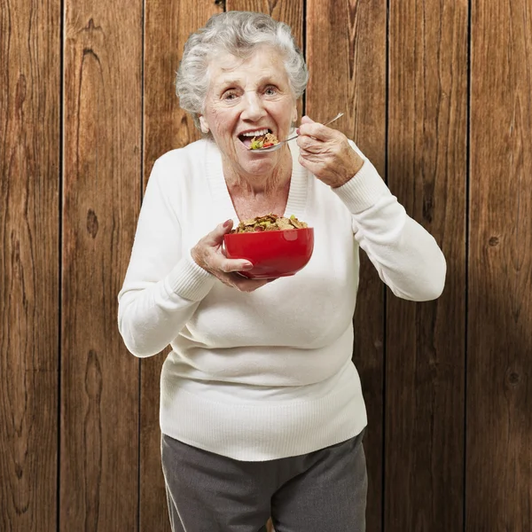 Senior woman eating cereals out of a red bowl against a wooden b — Stock Photo, Image