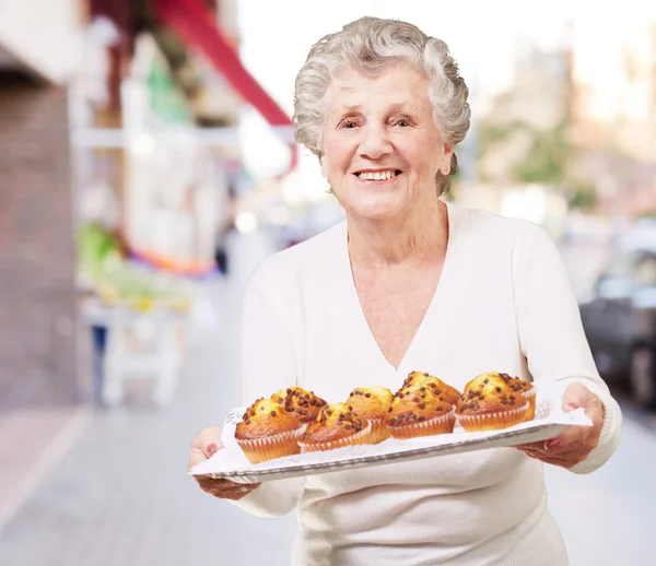 Retrato de una mujer mayor mostrando una bandeja de magdalenas de chocolate en stre — Foto de Stock