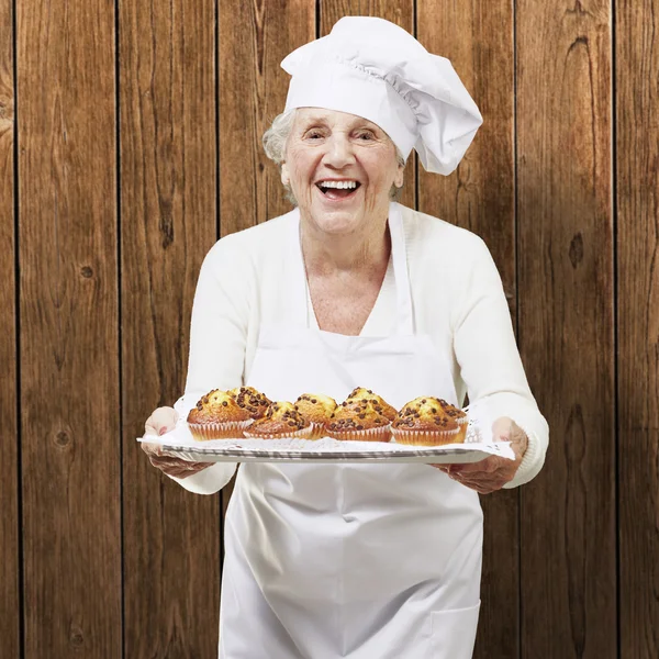 Senior woman cook holding a tray with muffins against a wooden b — Stock Photo, Image