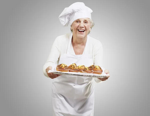 Senior woman cook holding a tray with muffins against a grey bac — Stock Photo, Image