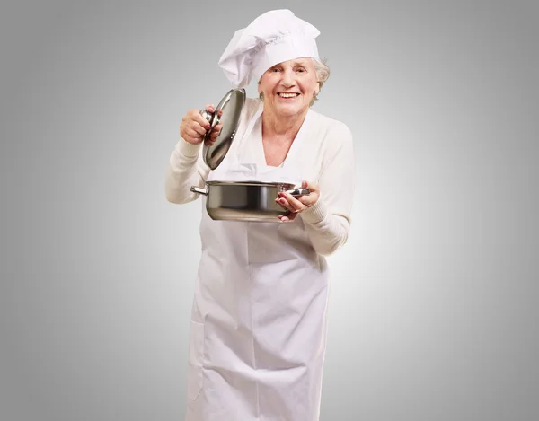 Портрет пожилой женщины, открывающей кастрюлю с соусом поверх серого — стоковое фото