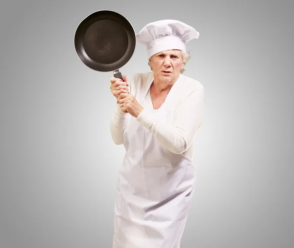 Cook ανώτερος γυναίκα θυμωμένος προσπαθεί να χτυπήσει με το τηγάνι πάνω από γκρι backgro — Φωτογραφία Αρχείου