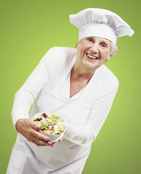 Cozinheiro de mulher sênior que mantém uma tigela com salada contra umas costas verdes — Fotografia de Stock