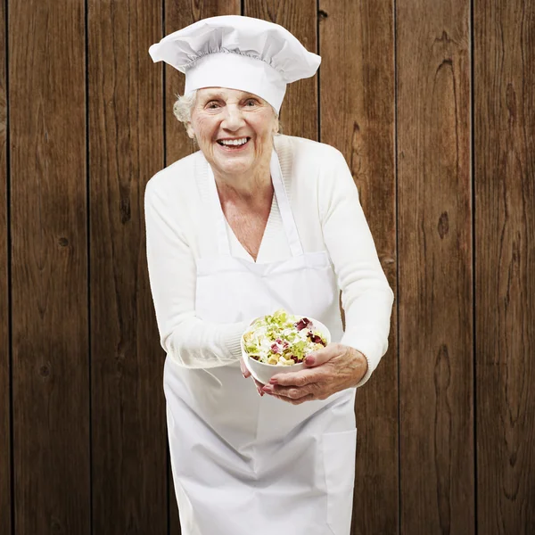 Mujer mayor cocinera sosteniendo un tazón con ensalada contra un bac de madera — Foto de Stock