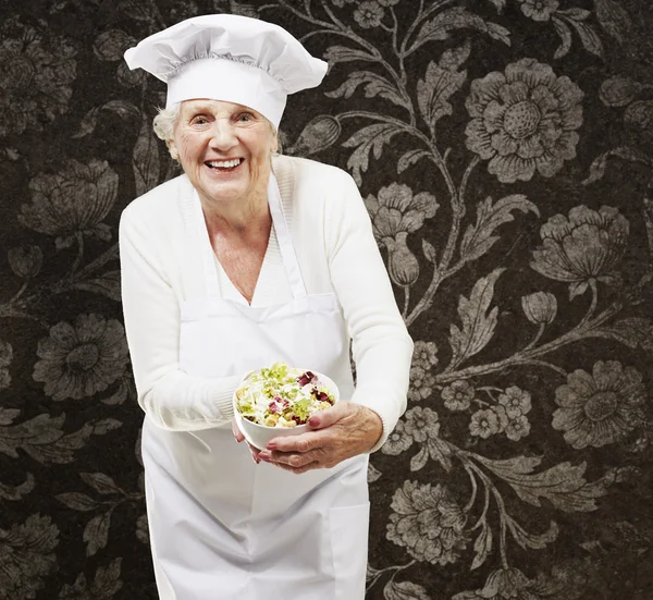Mujer mayor cocinera sosteniendo un tazón con ensalada contra una ba vintage —  Fotos de Stock