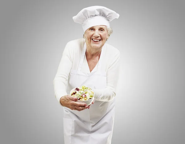 Mujer mayor cocinera sosteniendo un tazón con ensalada contra un respaldo gris — Foto de Stock