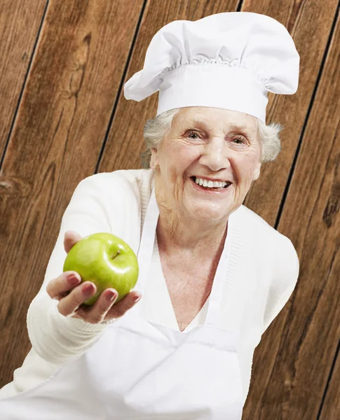 Senior woman cook offering a green apple against a wooden backgr — Stok fotoğraf