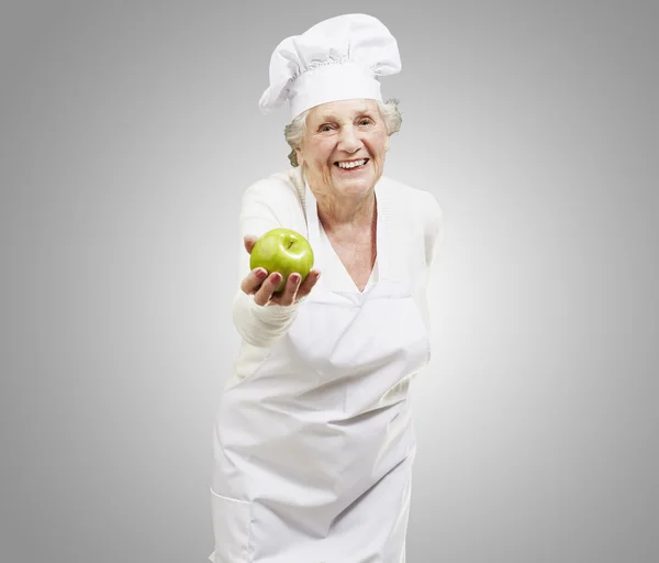 Mujer mayor cocinera ofreciendo una manzana verde contra un fondo gris —  Fotos de Stock