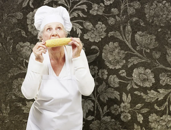 Senior woman cook eating a corncob against a vintage background — Stock Photo, Image