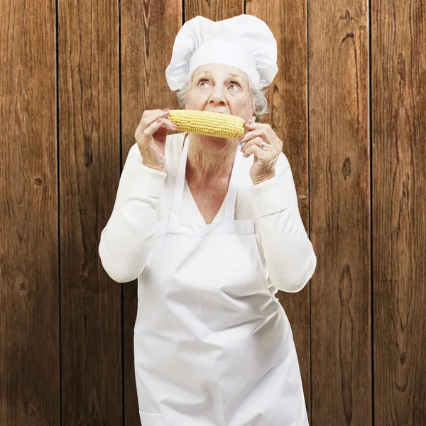 Senior woman cook eating a corncob against a wooden background — Stock Photo, Image