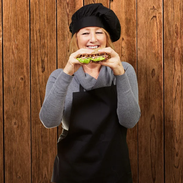 Middle aged cook woman holding a vegetal sandwich against a wood — Stock Photo, Image