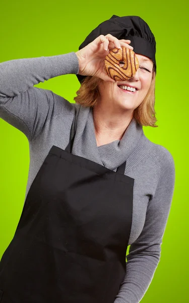 Middle aged cook woman looking through a donut over green backgr — Stock Photo, Image