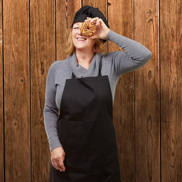 Middle aged cook woman looking through a donut against a wooden — Stock Photo, Image