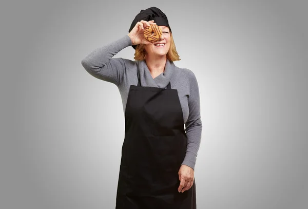 Középkorú cook nő nézegette egy fánkot, mint szürke backgro — Stock Fotó