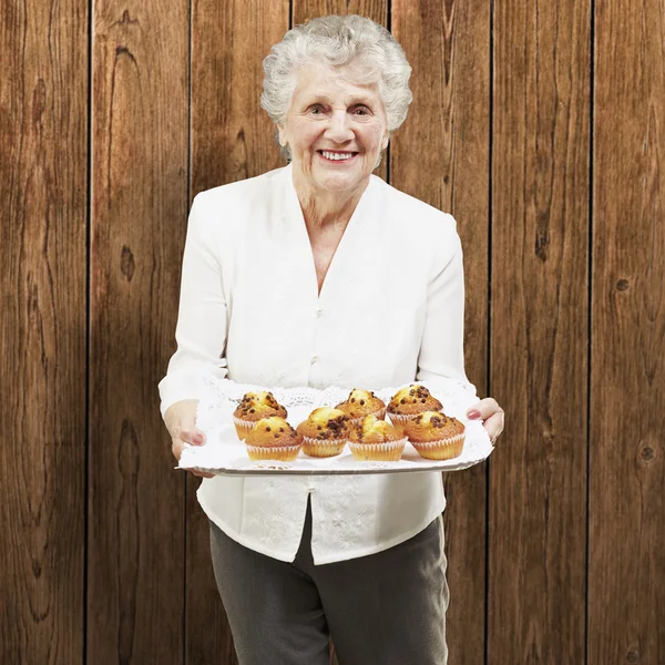 Seniorin lächelt und hält Tablett mit Muffins gegen eine Wand — Stockfoto
