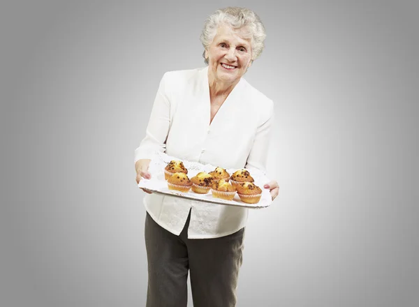 Senior woman smiling and holding a tray with muffins against a g — Stock Photo, Image