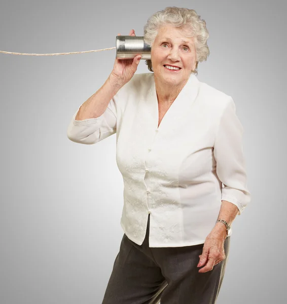Portrait of senior woman hearing with metal tin can over grey ba — Stock Photo, Image