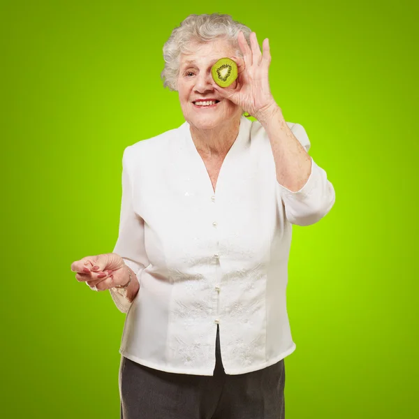 Portrait of senior woman holding kiwi in front of her eye over g — Stock Photo, Image