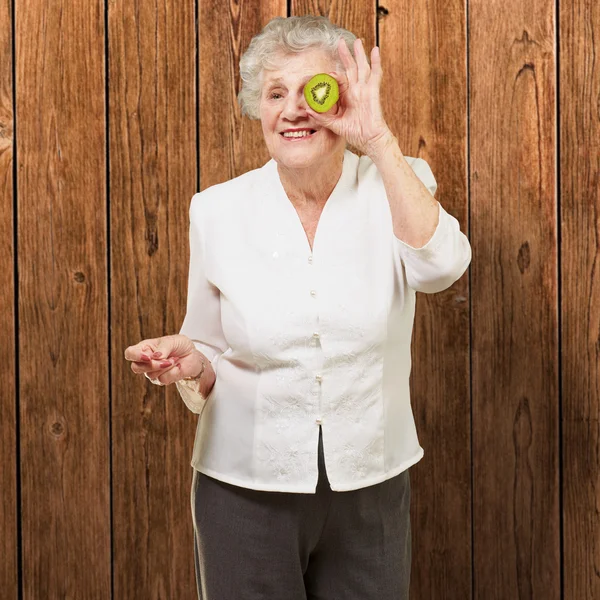 Portrait of senior woman holding kiwi in front of her eye agains — Stock Photo, Image